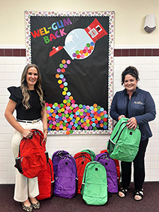 Harry Hoag Elementary school principal Amy Bartholomew and a representative of Hill & Markes, who are looking at and smiling for the camera, hold and stand in front of a row of colorful backpacks.