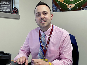 Fort Plain CSD Jr/Sr HIgh School Principal, Nicholas Comproski, who has short dark brown hair and is wearing a pink dress shirt and patterned tie, looks at and smiles for the camera while seated at a work desk. 