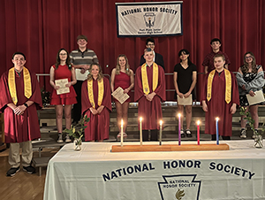A group of high school students stand together on risers on a stage in front of a dark red curtain. They are wearing long dark red National Honor Society robes and yellow drapes. They are looking at and smiling for the camera.