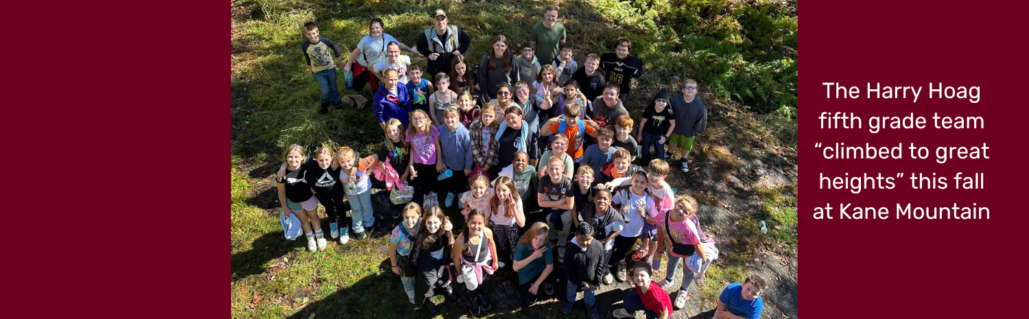 Harry Hoag fifth graders and teachers look up and smile for the camera in this aerial photo. The are all wearing hiking clothing and stand on green grass with rocky patches. The image features the phrase "The Harry Hoag fifth grade team climbed to great heights at Kane Mountain."