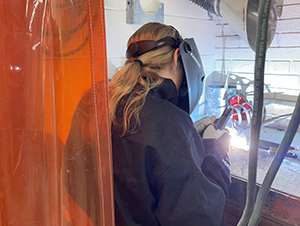 Sparks fly as a high school student wearing protective clothing and a welder's helmet welds in a school classroom.